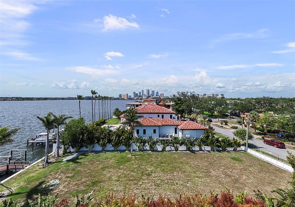 This aerial view showcases a waterfront property with a well-manicured lawn, a modern house with a red tile roof, and a private dock. The property is surrounded by lush landscaping and a white fence, offering both privacy and curb appeal. In the background, a cityscape adds to the property's desirable location.