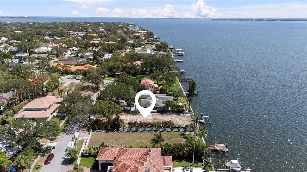 This aerial shot showcases a waterfront property with a house and a vacant lot marked by a location pin. The property features a well-maintained lawn, mature trees, and a private dock extending into the water. The surrounding neighborhood consists of similar upscale homes, and the overall impression is one of luxury and exclusivity.