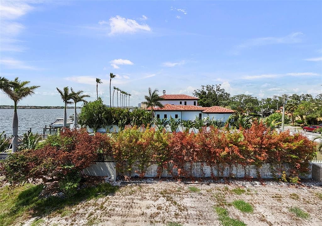 This aerial view showcases a luxurious waterfront property with a Mediterranean-style home featuring a red tile roof and white exterior walls. Lush landscaping surrounds the property, including palm trees and vibrant bushes, enhancing its curb appeal. The property also includes a dock on the water, suggesting waterfront access and recreational opportunities.
