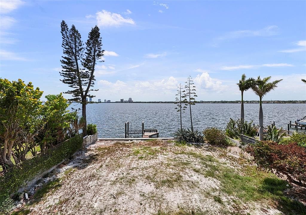 This exterior rear view showcases a waterfront property with a sandy yard leading to a calm body of water. A small dock extends into the water, and the skyline of a distant city is visible on the horizon. Lush greenery and palm trees frame the scene, creating a serene and inviting atmosphere.