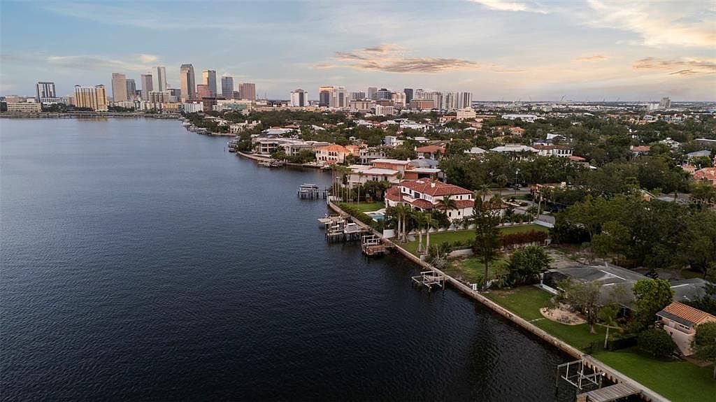 This aerial view showcases a waterfront residential area with luxurious homes, each featuring private docks and manicured lawns. The cityscape is visible in the background, creating a stunning contrast between urban and suburban living. The calm water reflects the sky, adding to the serene and upscale atmosphere of the neighborhood.