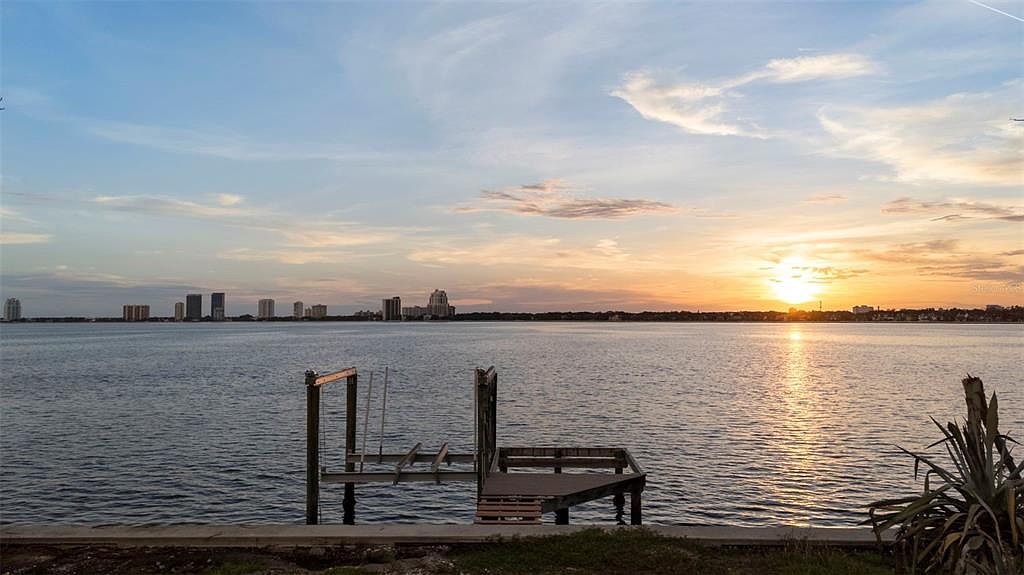 This exterior shot showcases a serene waterfront view at sunset, featuring a small wooden dock extending into the water. The cityscape is visible in the distance, adding an urban touch to the natural scenery. The overall impression is tranquil and inviting, highlighting the property's desirable location and potential for relaxation.