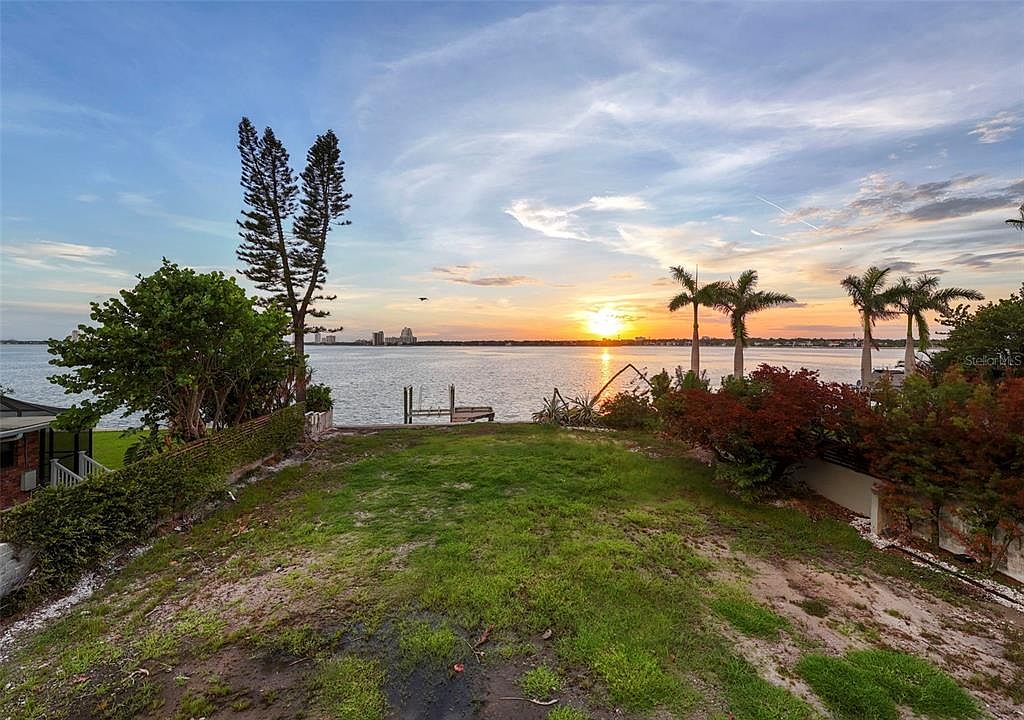 This image showcases a waterfront property's yard at sunset. The grassy area slopes down towards a calm body of water, with a small dock visible. Palm trees and other greenery line the waterfront, and the setting sun casts a warm glow over the scene, creating a serene and inviting atmosphere.