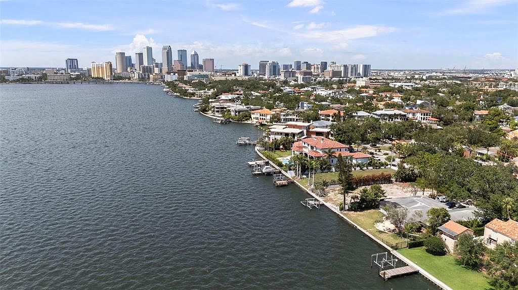 This aerial shot showcases a waterfront property with a stunning view of the city skyline. The luxurious homes along the shoreline feature well-manicured lawns, private docks, and lush landscaping. The calm water reflects the sky, creating a serene and upscale atmosphere, perfect for highlighting the property's prime location and exclusive lifestyle.
