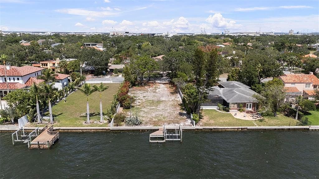 This aerial shot showcases a waterfront property featuring a cleared lot ready for construction, flanked by well-maintained homes. The property includes a private dock extending into the water, suggesting direct access for boating or other water activities. Lush greenery surrounds the area, providing a serene and upscale residential setting.