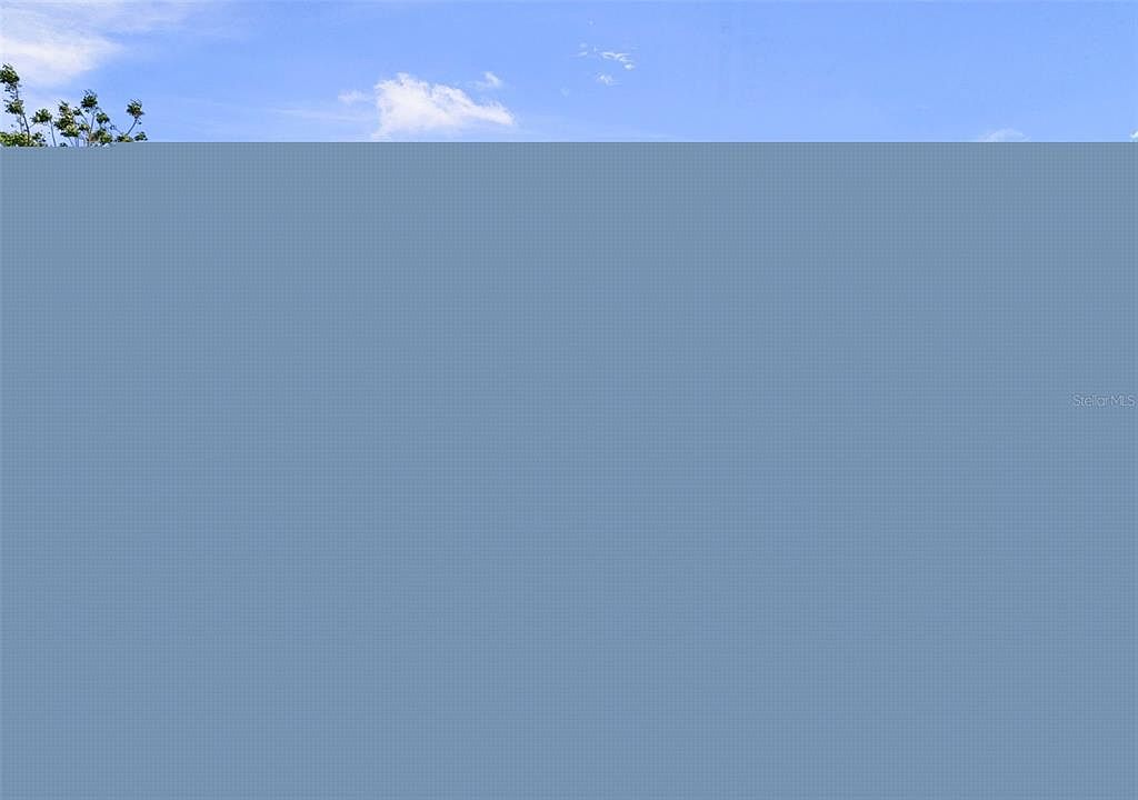 The image presents an aerial view of a clear blue sky with scattered clouds. A few treetops are visible in the upper left corner. The overall impression is serene and expansive, suggesting a property with open space and pleasant weather conditions.