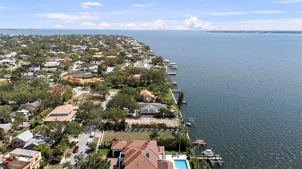 This aerial view showcases a waterfront property with a luxurious home featuring a red tile roof and a private pool. The property includes a well-maintained yard and a dock extending into the water, suggesting direct access for boating. The surrounding neighborhood is lush with mature trees and upscale homes, emphasizing the desirable location.