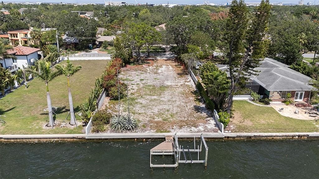 This aerial shot showcases a waterfront property featuring a vacant lot with a dock and boat lift. The lot is bordered by well-maintained landscaping and a seawall. Adjacent to the lot is a single-story home with a screened-in porch and mature trees, offering a glimpse of the neighborhood beyond.