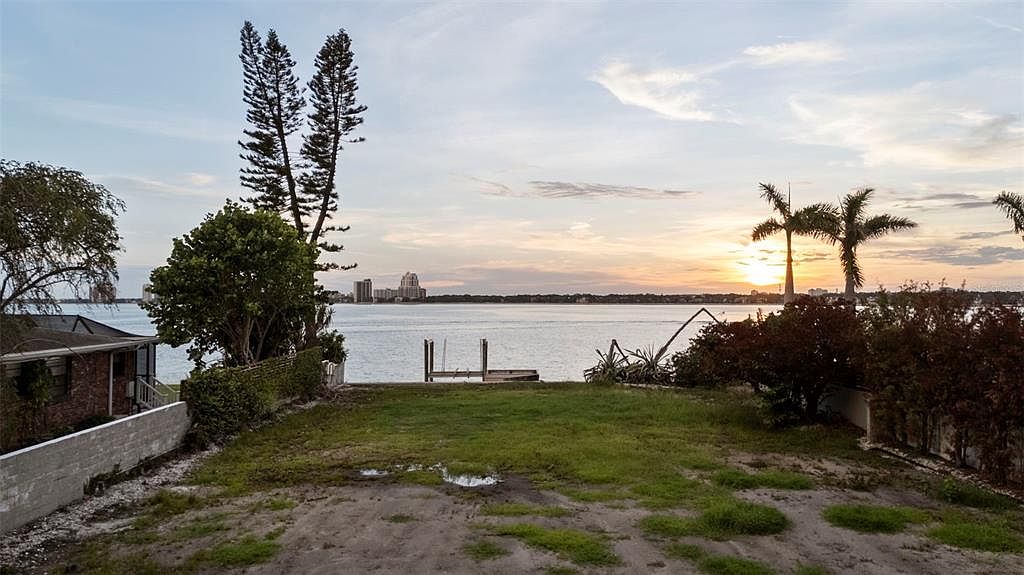 This exterior rear view showcases a waterfront property with a gently sloping yard leading to the water. A small dock is visible, and the cityscape can be seen in the distance during sunset. The yard is a mix of grass and sandy areas, bordered by trees and shrubs, offering a serene and private setting.