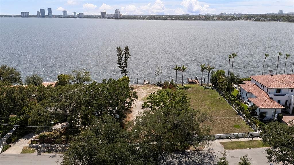 This aerial view showcases a waterfront property featuring a vacant lot with potential for development. The lot is partially cleared with some grass and trees, leading to a waterfront with docks. A large, luxurious house with a red tile roof is visible on the adjacent property, and the cityscape is visible in the background across the water.