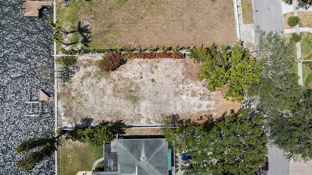 This aerial view showcases a vacant lot situated between a waterfront and a residential street. The lot appears cleared and ready for development, with some mature trees along the property lines. A dock extends into the water, suggesting potential for waterfront living.