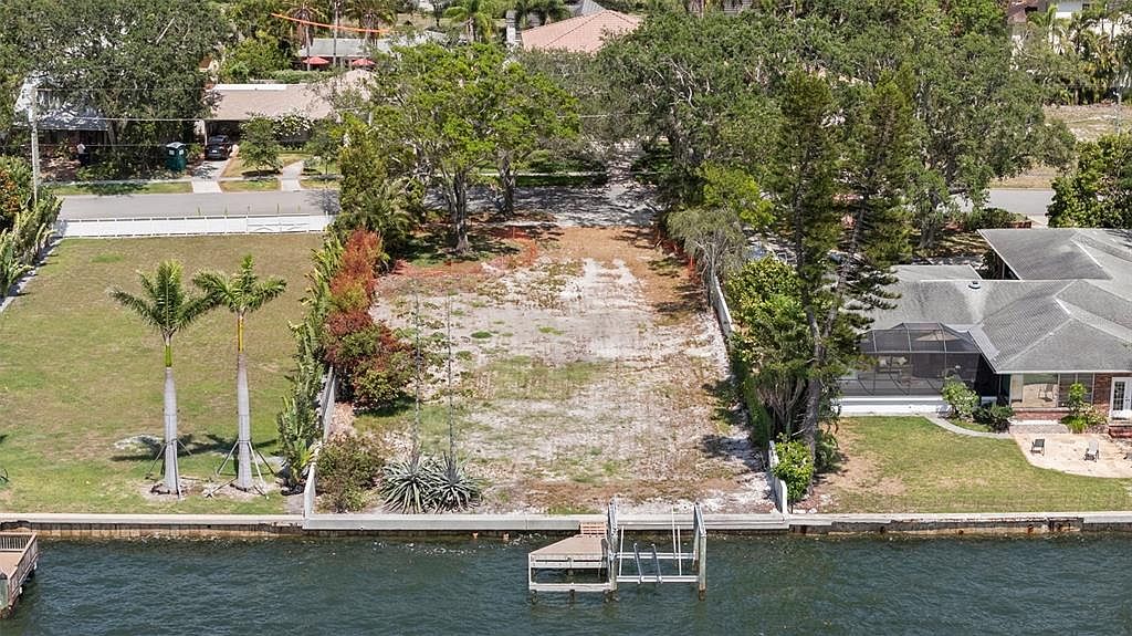 This aerial shot showcases a waterfront property, highlighting a vacant lot with a private dock extending into the water. The lot is bordered by mature trees and landscaping, with neighboring houses visible on either side. The image emphasizes the property's waterfront location and potential for development.
