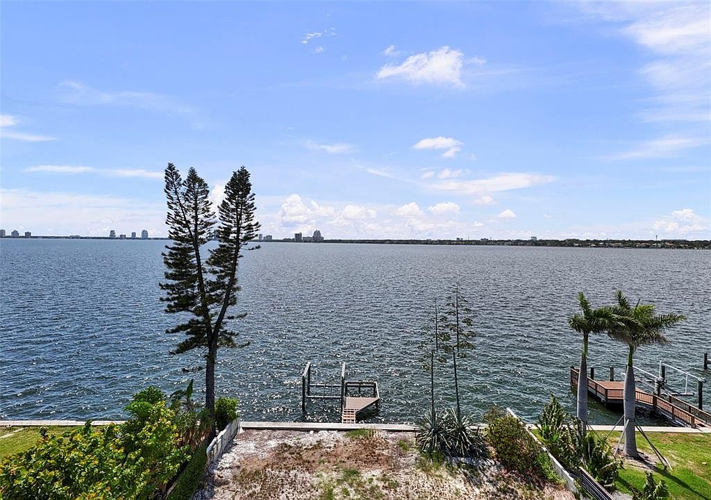 This exterior rear view showcases a waterfront property with a serene ocean backdrop under a partly cloudy sky. The property features a small pier extending into the water, along with mature trees and landscaping along the shoreline. The view emphasizes the property's access to the water and its potential for outdoor enjoyment.