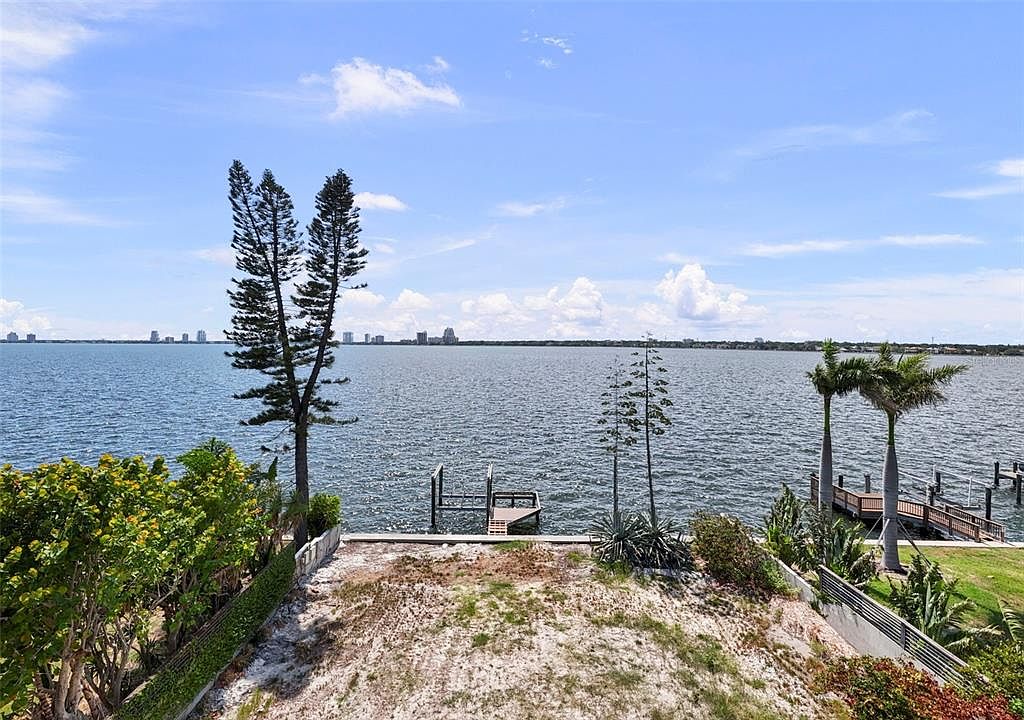 This exterior shot showcases the rear of a waterfront property, featuring a sandy yard leading to a calm bay. A small dock extends into the water, and mature trees and landscaping frame the view. The cityscape is visible in the distance, adding to the property's appeal.