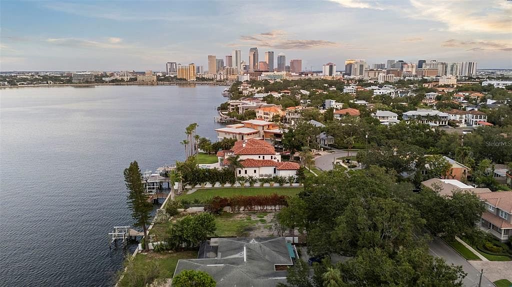 This aerial view showcases a luxurious waterfront property with a red-tiled roof, lush green lawn, and private dock. The home is nestled among mature trees, offering both privacy and stunning views of the bay and the city skyline in the distance. The overall impression is one of upscale coastal living.