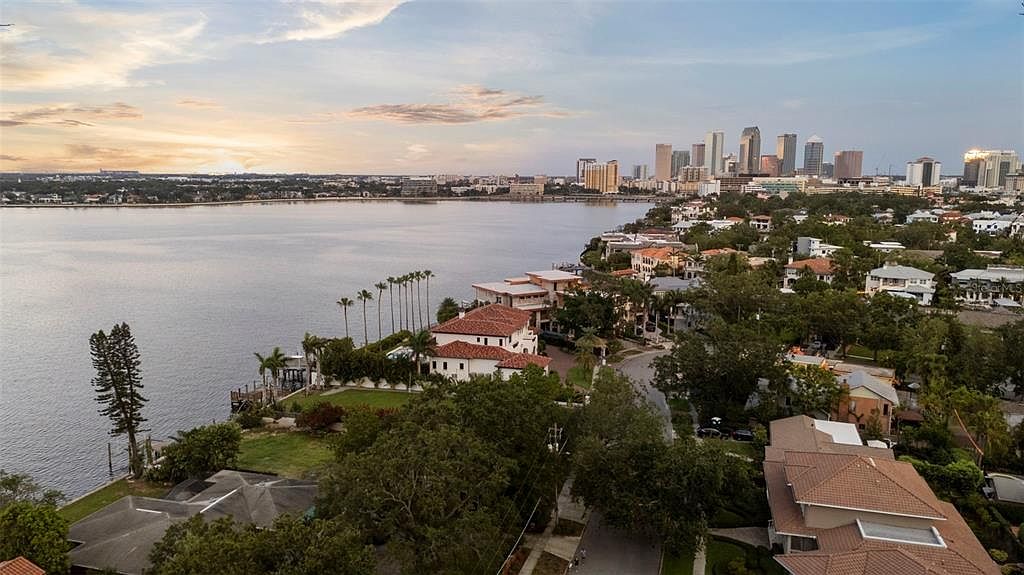 This aerial view showcases a luxurious waterfront property with a red-tiled roof and lush landscaping. The home is situated on a bay, offering stunning views of the water and the city skyline in the distance. The overall impression is one of exclusivity and high-end living.