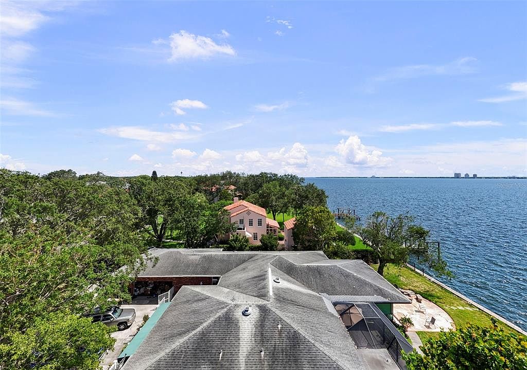 This aerial shot showcases a sprawling residential property with a dark gray roof, lush green landscaping, and waterfront access. A secondary building with a red tile roof is nestled among the trees. The view extends to a calm body of water, suggesting a serene and private setting.
