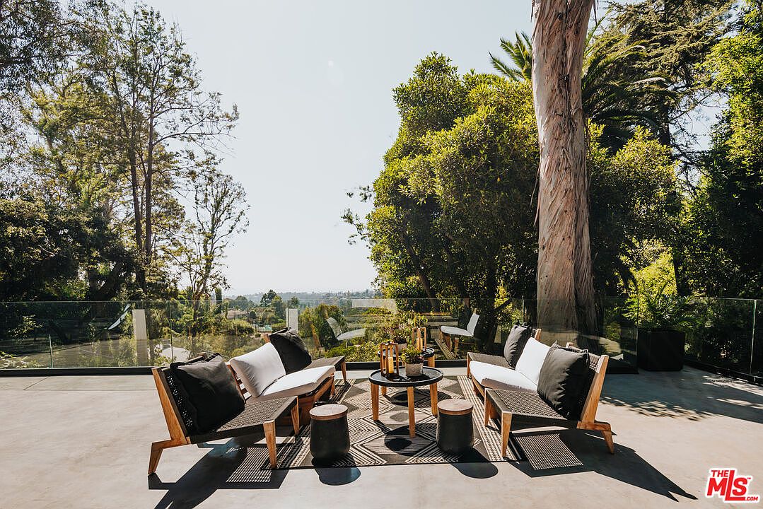 This image showcases a stylish outdoor patio area, featuring modern lounge furniture arranged around a low coffee table. The seating area is defined by a patterned rug and complemented by small side tables. A glass railing provides an unobstructed view of the surrounding landscape, blending the outdoor living space seamlessly with nature.