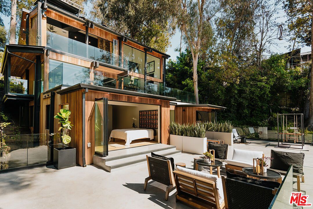 This image showcases a modern home's outdoor patio area, featuring stylish patio furniture arranged on a concrete surface. The patio seamlessly connects to an open bedroom through large glass doors, offering an indoor-outdoor living experience. Lush greenery surrounds the patio, enhancing the sense of privacy and tranquility.