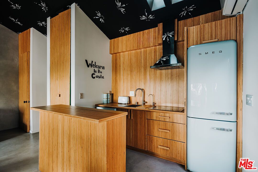 This is a modern kitchen with light wood cabinetry and a light blue SMEG refrigerator. The kitchen features a black range hood and a small island. The ceiling is black with white dragonfly patterns, adding a unique design element to the space.