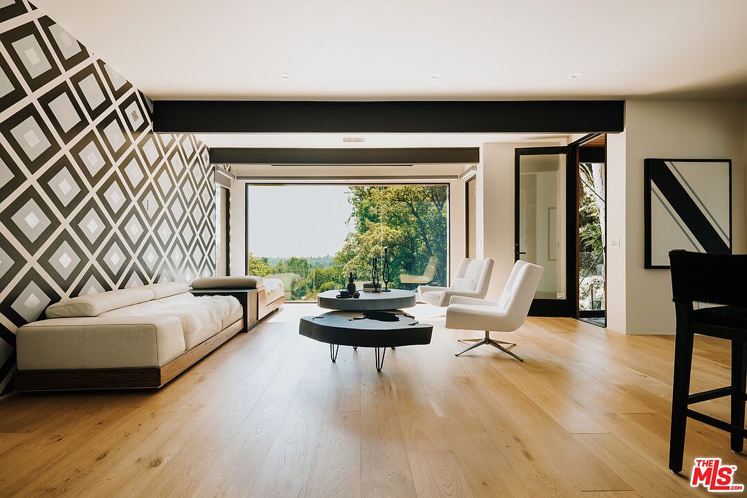 This is a bright and modern living room featuring hardwood floors, a geometric patterned accent wall, and a large window offering a view of lush greenery. The room is furnished with a contemporary sofa, unique coffee tables, and stylish white armchairs, creating an inviting and sophisticated atmosphere. The perspective is from the corner of the room, showcasing the open layout and connection to the outdoors.