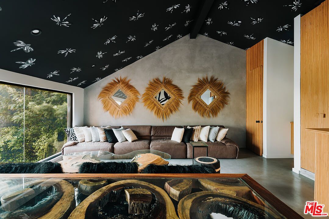 This interior shot showcases a stylish living room with a unique aesthetic. The room features a large brown sectional sofa adorned with various pillows, three decorative straw-framed mirrors on the wall, and a distinctive coffee table made of wood and glass. The ceiling is painted black with white dragonfly patterns, adding a whimsical touch to the space.