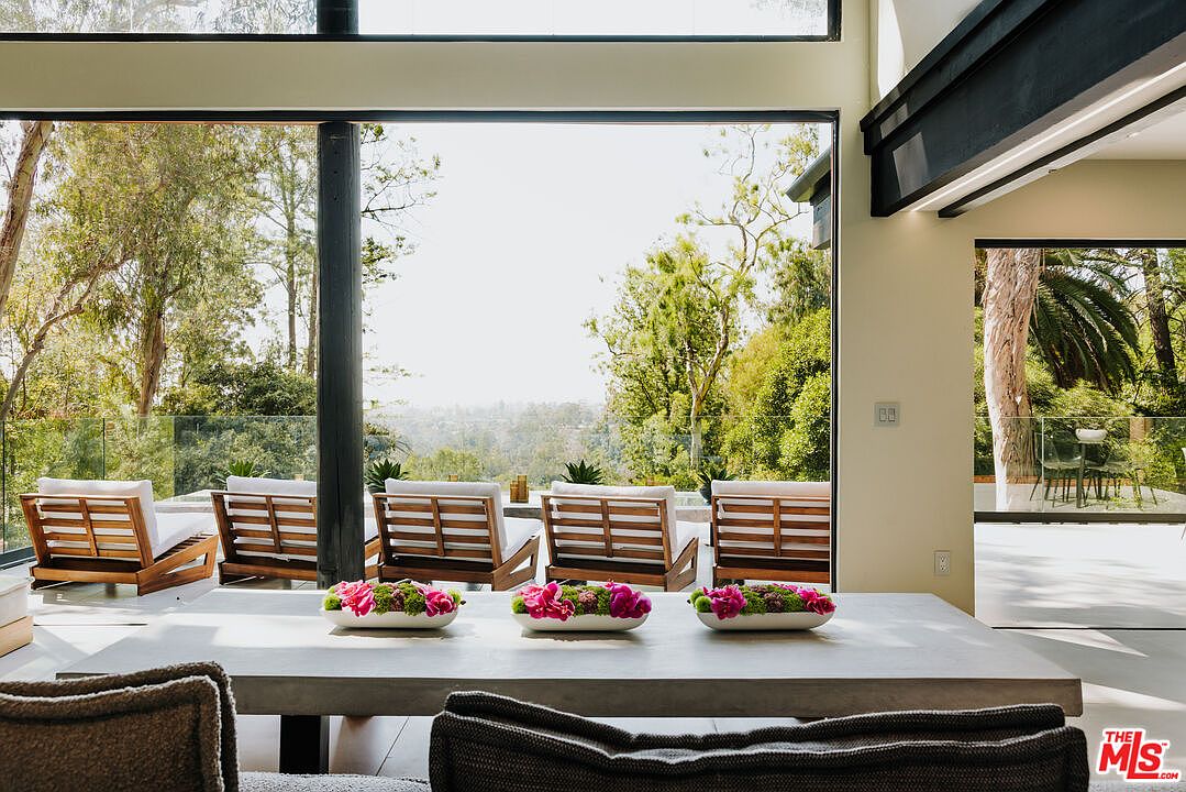 This image showcases a modern dining room with a seamless transition to an outdoor patio. The dining area features a large concrete table adorned with floral centerpieces, complemented by stylish chairs. Expansive glass doors offer unobstructed views of lush greenery and a distant cityscape, creating an open and airy atmosphere.