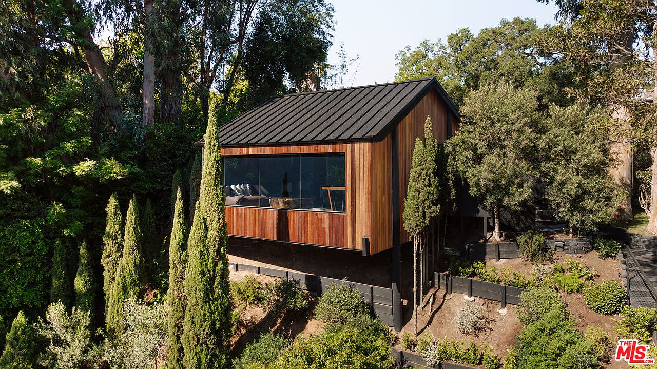 This image showcases a modern, elevated cabin-style home nestled among lush greenery. The structure features a striking combination of dark metal roofing and warm wood siding, with a large picture window offering a glimpse into the interior. The surrounding landscape includes mature trees and manicured shrubs, creating a serene and private setting.