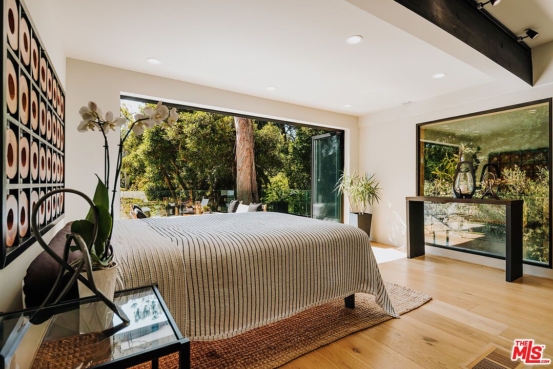 This is a bright and airy primary bedroom featuring hardwood floors, a large bed with a striped cover, and a wall of windows overlooking a lush outdoor area. The room is decorated with modern art and plants, creating a serene and inviting atmosphere. A large mirror on a console table reflects the outdoor scenery, enhancing the sense of space and connection to nature.