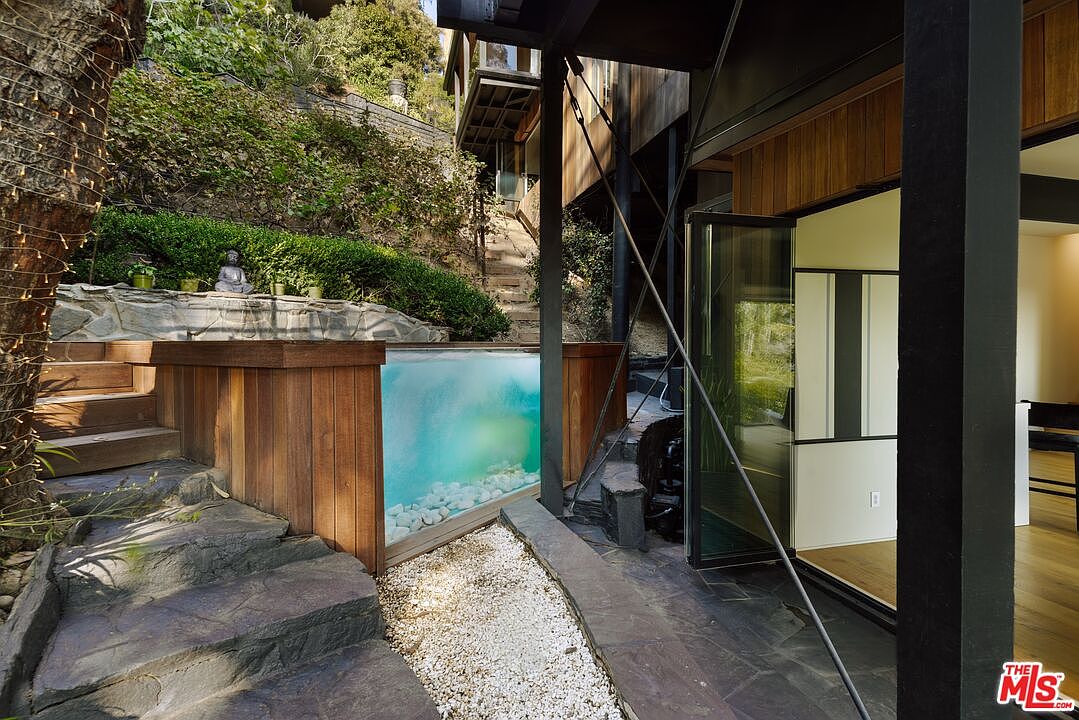 This image showcases a unique pool/spa feature integrated into the landscape of a modern home. The pool has a glass side, offering a view into the water, and is surrounded by wooden decking and stone steps. The area is landscaped with lush greenery, creating a tranquil and visually appealing outdoor space.