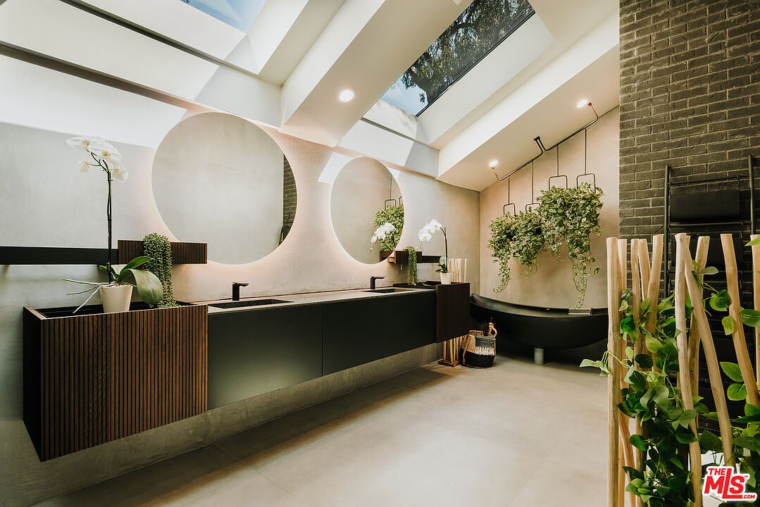 This is a modern primary bathroom featuring a double vanity with round mirrors and sleek black fixtures. The room is well-lit with natural light from skylights and accented with hanging plants, creating a spa-like atmosphere. A freestanding tub is visible in the background, and the overall design incorporates natural elements and a minimalist aesthetic.