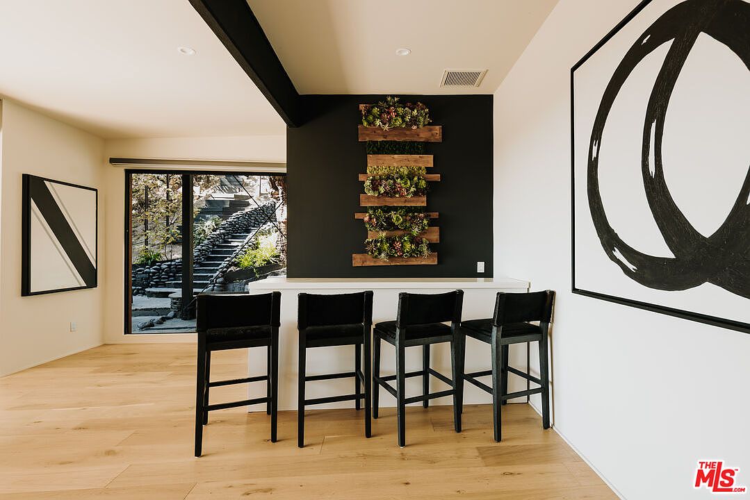 This interior shot showcases a modern living space with light wood flooring and a minimalist aesthetic. Four black bar stools are positioned at a white counter, set against a dark accent wall adorned with a vertical succulent display. Large abstract artwork hangs on the adjacent white wall, contributing to the room's sophisticated and contemporary feel.