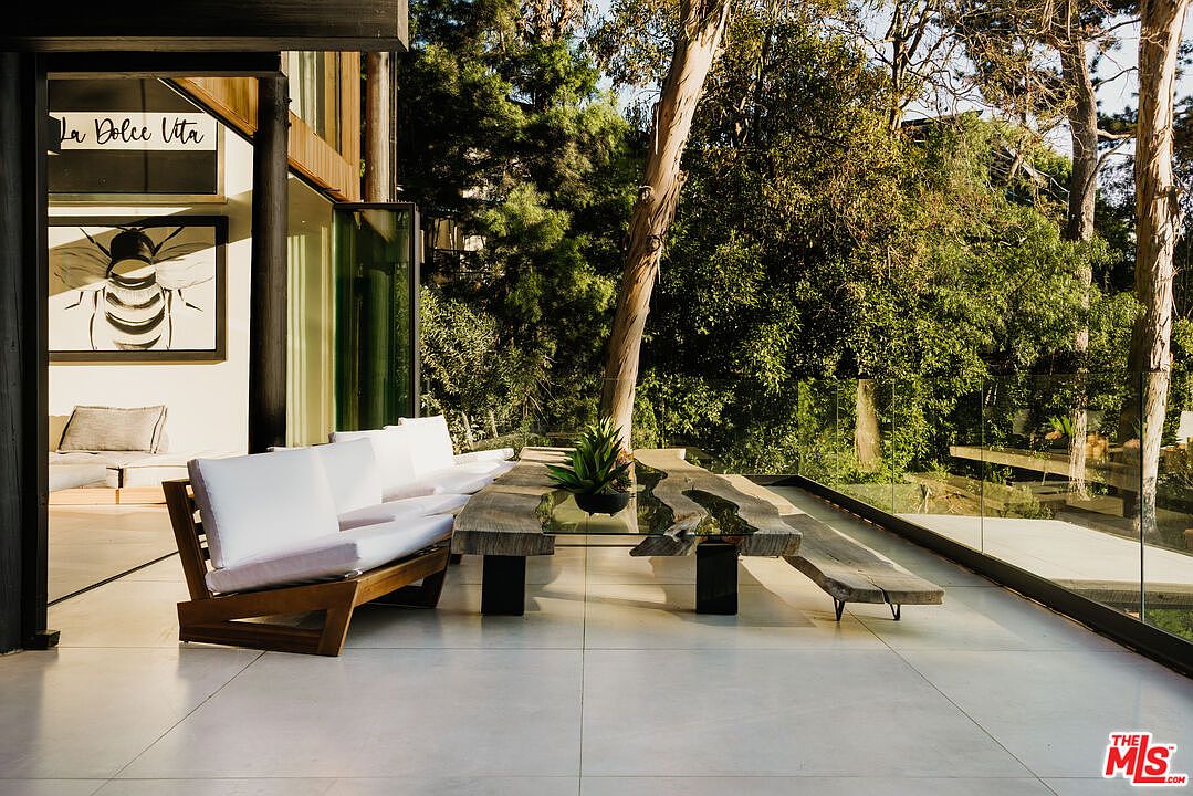 This image showcases a stylish outdoor patio area, featuring a unique wooden table with a glass insert and matching benches, complemented by a modern outdoor sofa with white cushions. The patio is surrounded by lush greenery and a glass railing, offering a seamless blend of indoor and outdoor living. The overall impression is one of sophisticated relaxation and connection with nature.