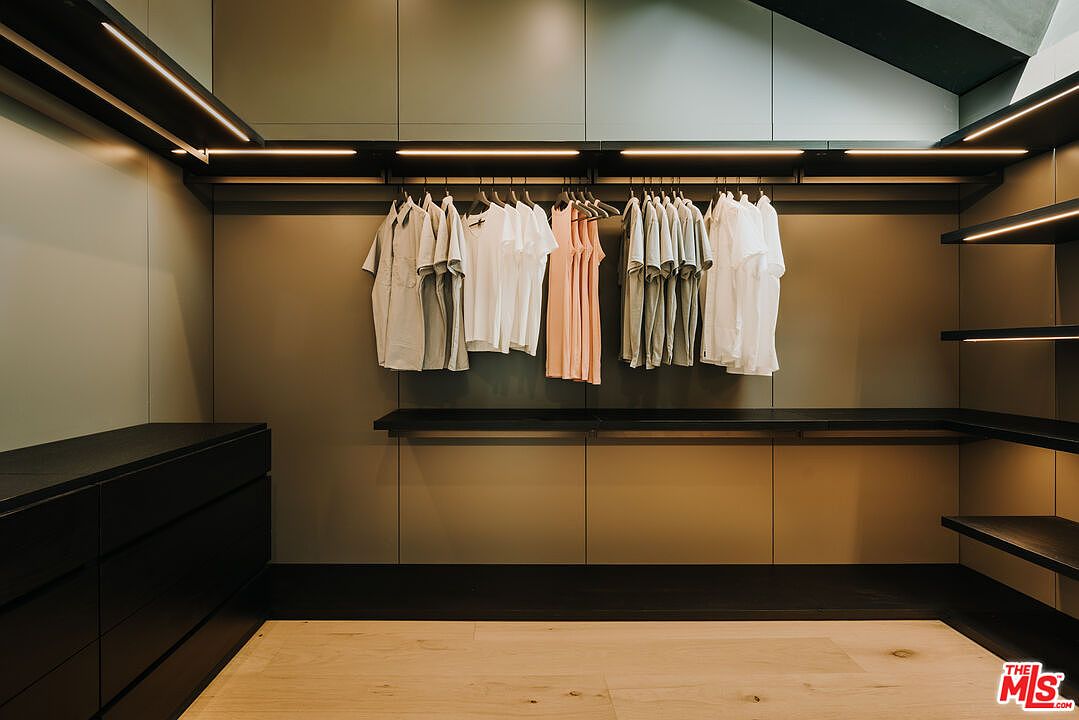 This is a well-organized walk-in closet featuring a variety of clothing items hanging neatly on rods, including shirts and dresses. The closet has built-in shelving and drawers, with integrated lighting that highlights the space. The overall design is modern and functional, providing ample storage.
