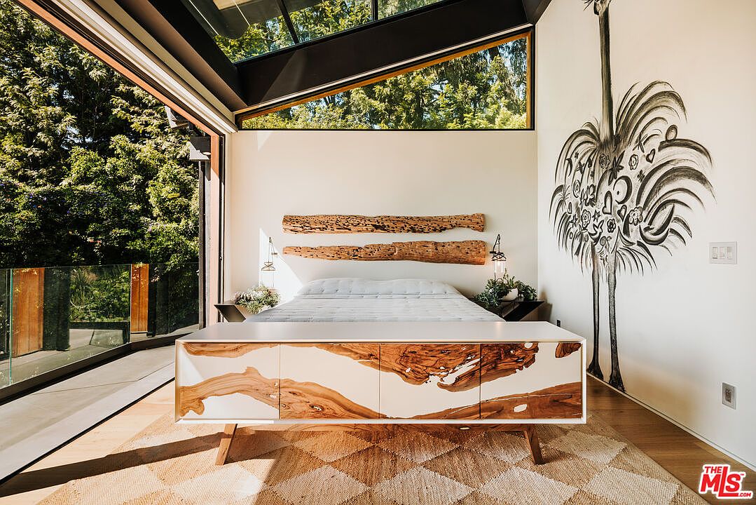 This is a bright and airy primary bedroom featuring a unique, rustic-modern design. The room boasts a custom wooden headboard and a matching console table with a white and wood grain finish. Large windows offer views of the surrounding greenery, and a whimsical ostrich mural adds a touch of artistic flair to the space.