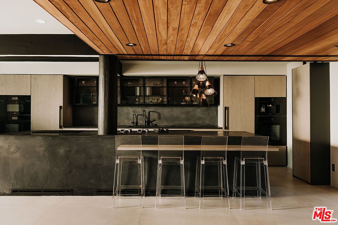 This is a modern kitchen with a unique wooden plank ceiling and a dark, textured island with seating. The cabinetry is a light wood tone, and there are built-in appliances. The kitchen features a sleek, minimalist design with a focus on natural materials and a neutral color palette.