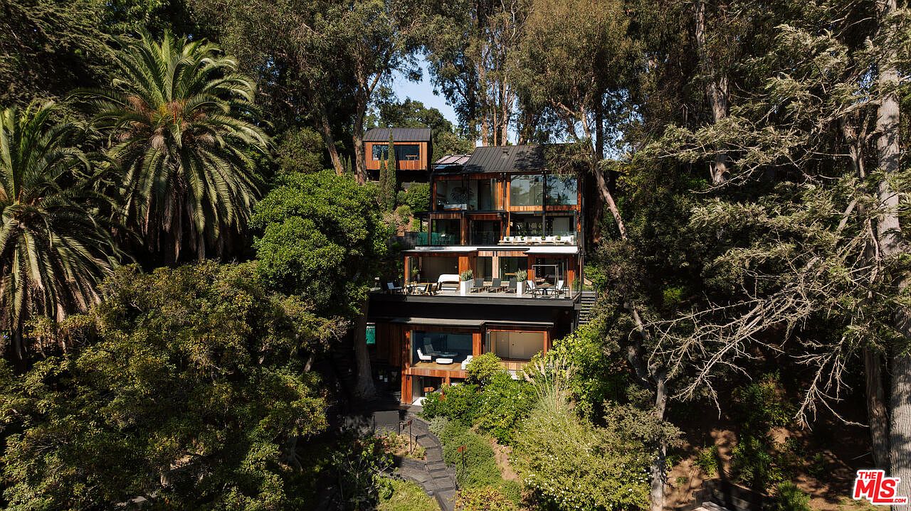 This aerial view showcases a modern, multi-level home nestled among lush trees and greenery. The architectural design features a combination of wood and glass, with multiple balconies and outdoor living spaces. A pathway leads to the house, emphasizing its integration with the natural surroundings.