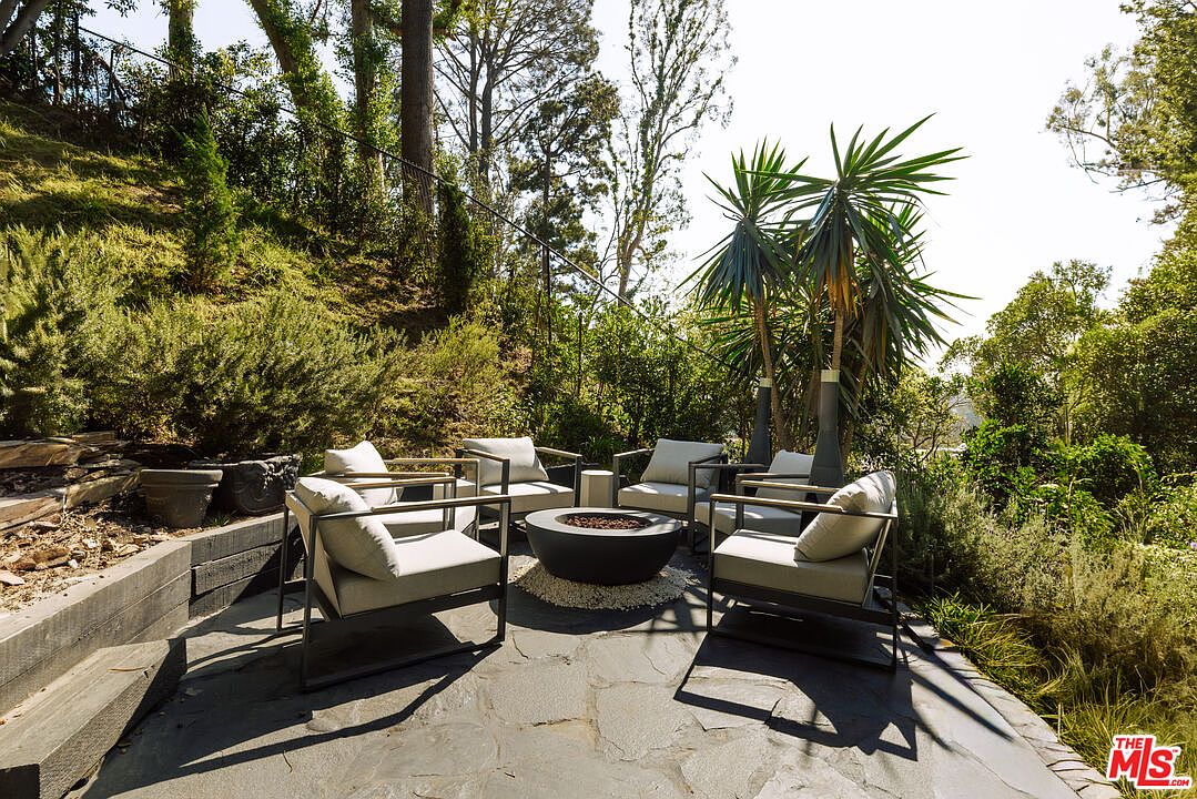 This image showcases a stylish outdoor patio area, featuring a circular fire pit surrounded by modern armchairs with light-colored cushions. The patio is paved with stone slabs and is nestled within lush greenery, creating a serene and inviting atmosphere. The view is from a medium distance, capturing the entire seating arrangement and the surrounding landscape.