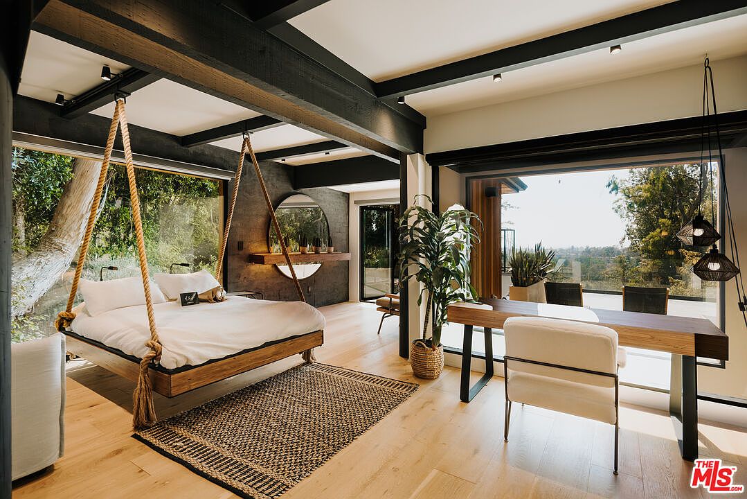 This primary bedroom features a unique hanging bed suspended by thick ropes, complemented by a textured area rug and light wood flooring. A modern desk area with a large window offers a view of the outdoors, while a round mirror and minimalist decor add to the room's contemporary style. The dark ceiling beams provide a striking contrast to the light walls and natural light flooding the space.
