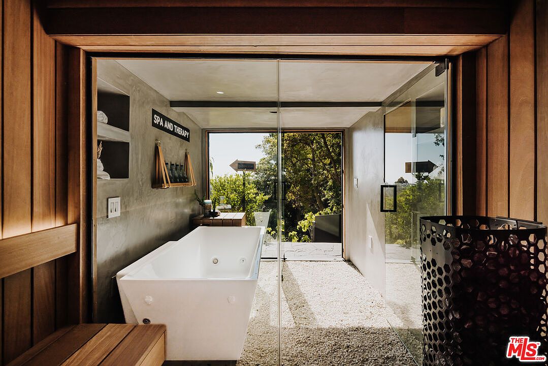 This image showcases a luxurious primary bathroom featuring a modern freestanding bathtub positioned near a glass-enclosed area with an outdoor view. The bathroom's design incorporates natural wood elements and a neutral color palette, creating a spa-like atmosphere. The perspective is from the doorway, inviting the viewer into the serene space.
