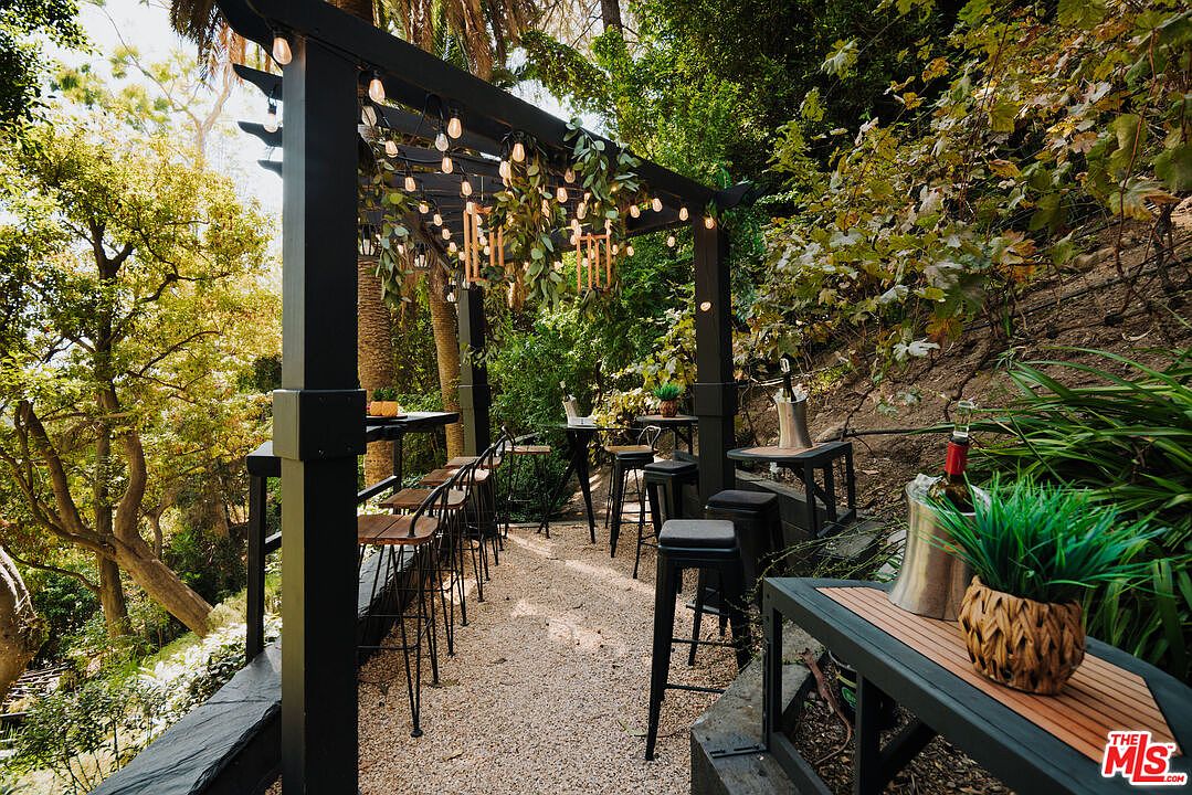 This image showcases an inviting outdoor patio area, featuring a dark pergola adorned with string lights and lush greenery. The space is furnished with high-top tables and bar stools, creating a relaxed and stylish setting for entertaining. The ground is covered with gravel, and the surrounding landscape includes trees and foliage, enhancing the natural ambiance.