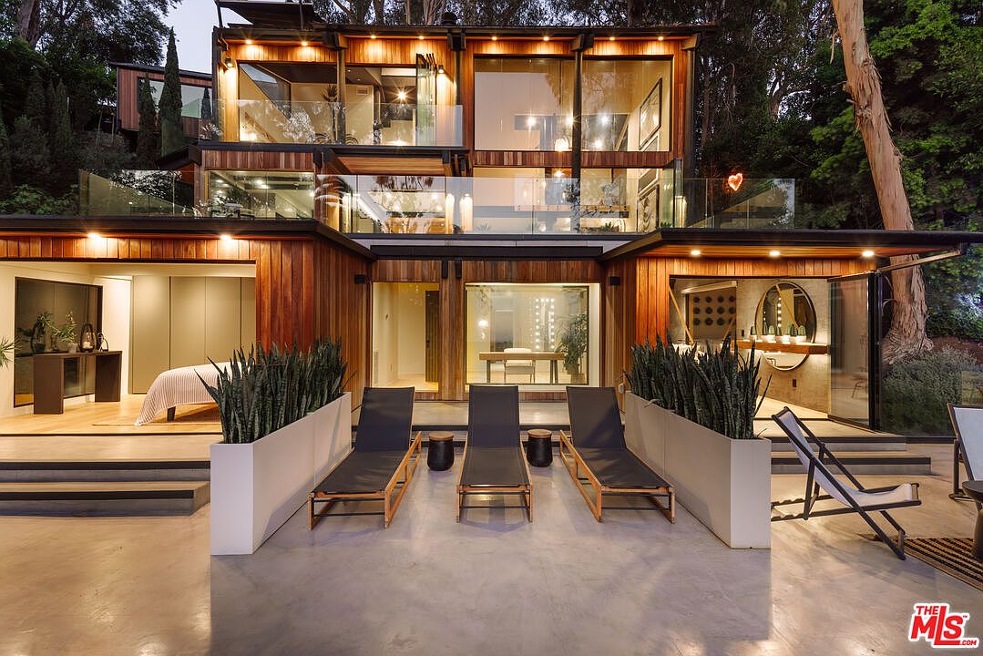 This is a rear exterior view of a modern, multi-level home featuring wood paneling and large glass windows. The outdoor space includes a patio with lounge chairs and planters, creating a relaxing atmosphere. The architecture emphasizes indoor-outdoor living with seamless transitions between the interior spaces and the exterior patio.