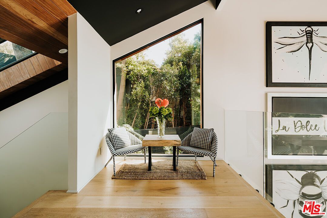 This interior shot showcases a stylish hallway or landing area with a modern aesthetic. A triangular window provides natural light and a view of lush greenery, while two houndstooth chairs and a small table create a cozy seating nook. The space is adorned with framed artwork and features light wood flooring, contributing to a bright and inviting atmosphere.