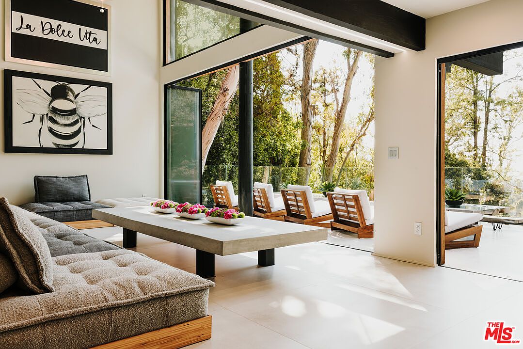 This is a bright and airy living room featuring a modern aesthetic with clean lines and neutral tones. The room opens to an outdoor patio area through large glass doors, seamlessly blending indoor and outdoor living. A large sectional sofa with tufted cushions and a concrete coffee table create a comfortable and stylish space.