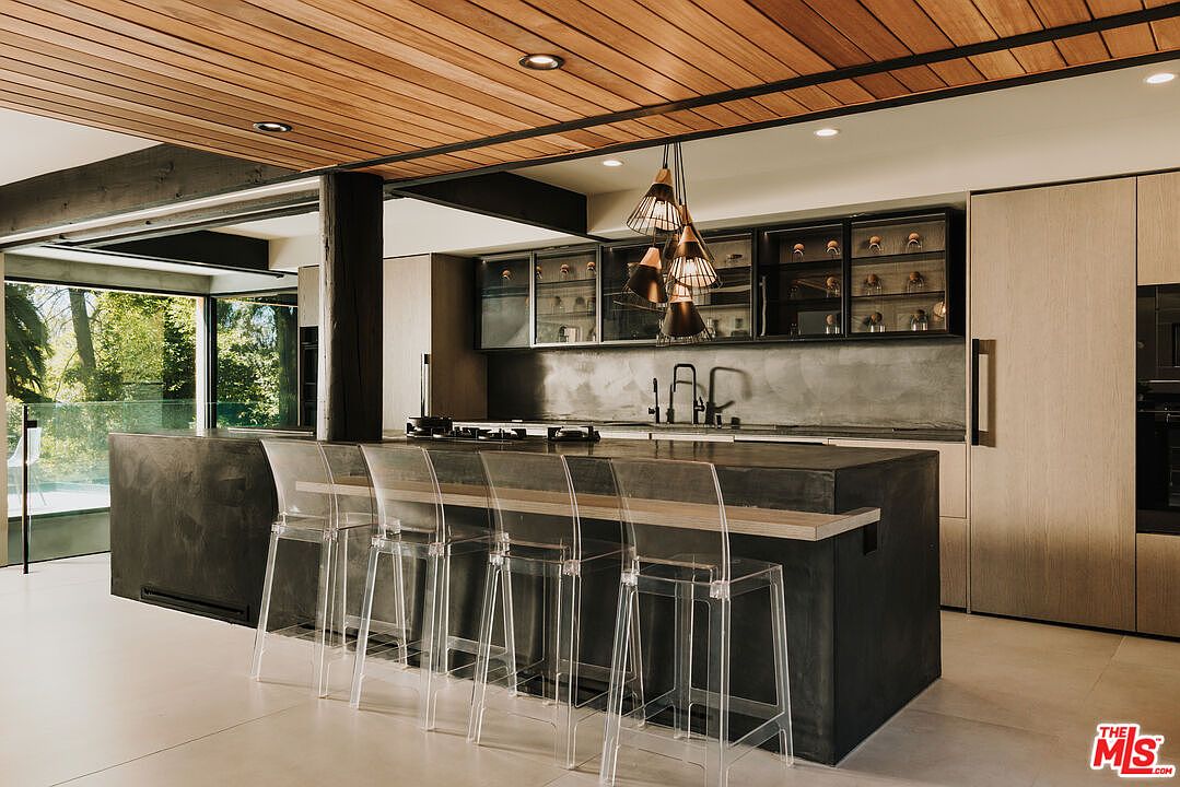 This is a modern kitchen with a large dark gray island featuring clear bar stools. The kitchen has sleek cabinetry, stainless steel appliances, and unique pendant lighting. A large window offers a view of the outdoors, and the ceiling is finished with wood panels.