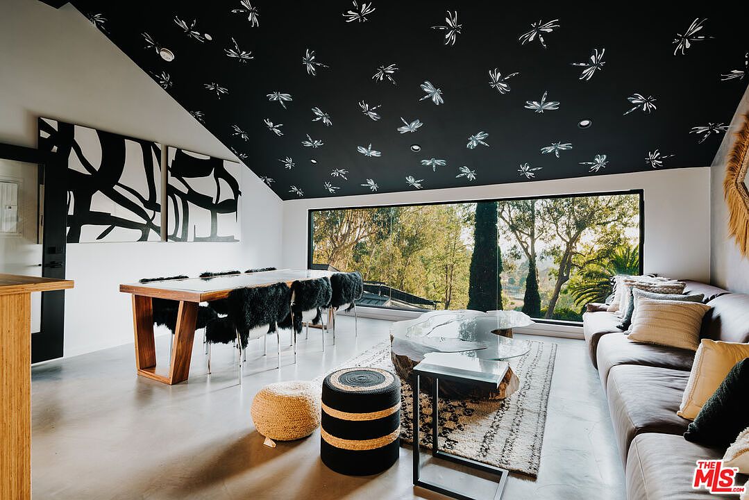 This interior shot showcases a modern living and dining area with a unique design. The room features a dining table with fur-covered chairs, a comfortable sofa with decorative pillows, and a stylish coffee table. The ceiling is adorned with a dragonfly pattern, adding a whimsical touch to the space, while large windows offer a view of the lush greenery outside.