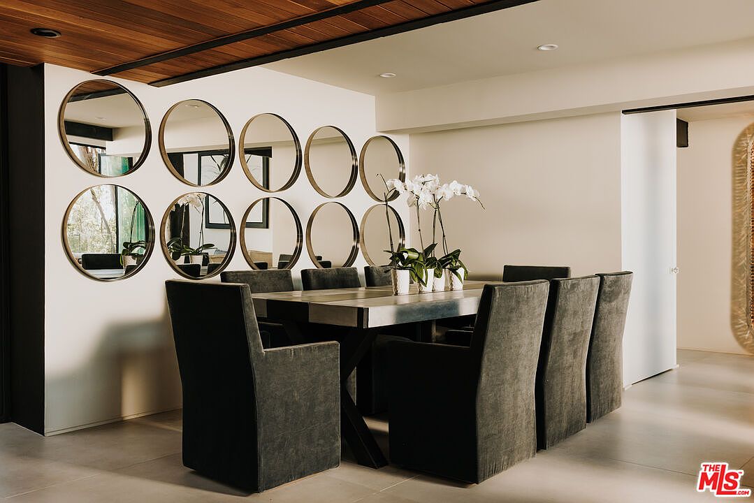 This interior shot showcases a modern dining room with a long table surrounded by upholstered chairs. A striking arrangement of round mirrors adorns the wall, reflecting light and adding visual interest. The room features a neutral color palette, complemented by a wood-paneled ceiling, creating a sophisticated and inviting atmosphere.