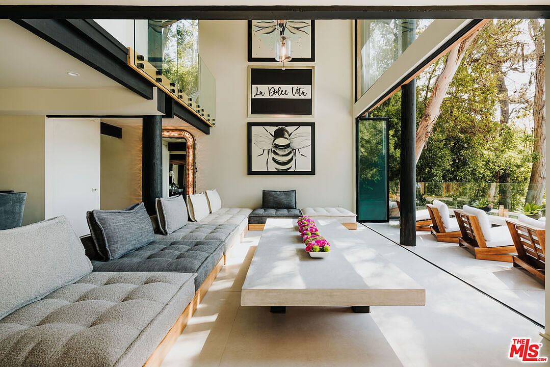 This is a bright and airy living room featuring a long, tufted sofa with gray cushions and a large, rectangular coffee table with a floral centerpiece. The room has floor-to-ceiling windows that offer a view of the outdoor patio and greenery, creating a seamless indoor-outdoor living experience. Artwork adorns the walls, and the overall style is modern and minimalist.