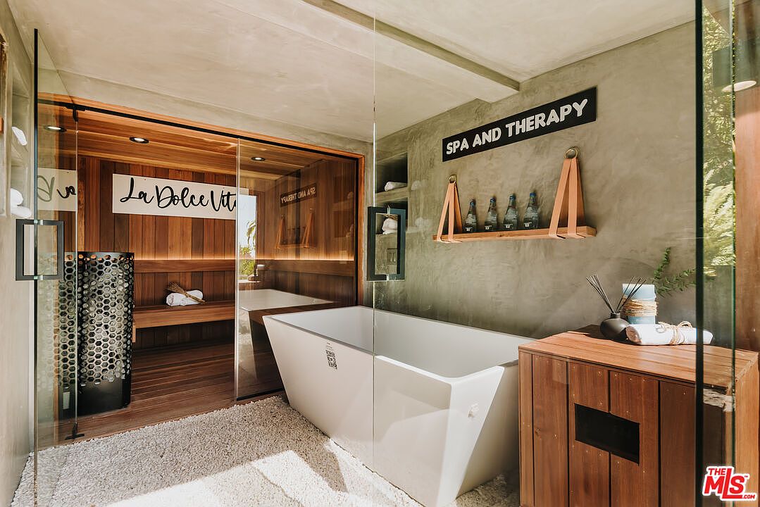 This primary bathroom showcases a modern design with a sauna, a freestanding bathtub, and a wooden vanity. The sauna features wood paneling and glass doors, while the bathtub is positioned against a textured gray wall with a decorative shelf. The overall aesthetic is luxurious and spa-like, creating a relaxing atmosphere.