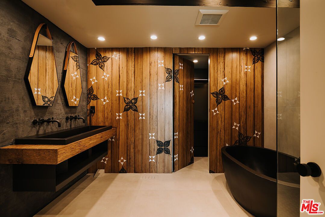 This is a modern primary bathroom featuring a unique wood-paneled wall with decorative floral patterns, a black vessel sink on a wooden vanity, and a freestanding black bathtub. The room is illuminated by recessed lighting, and the overall design creates a luxurious and spa-like atmosphere with a blend of rustic and contemporary elements.